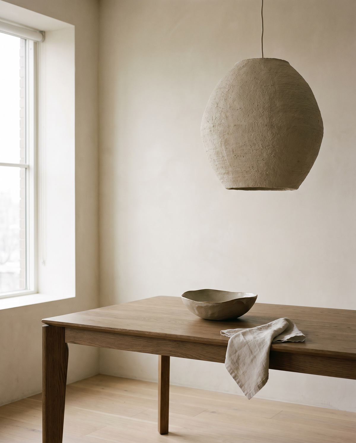 An oak dining table beneath a single pendant light in a calm ivory-walled room